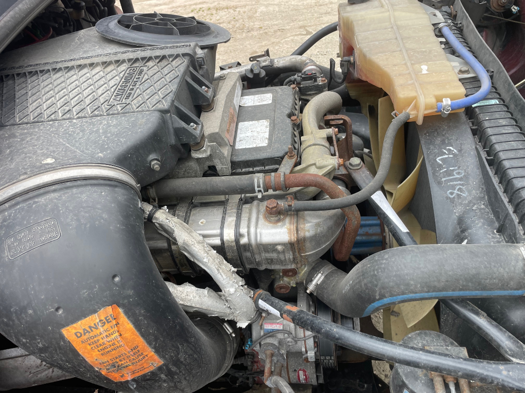 Truck engine bay of a 2014 Freightliner Cascadia 125 showing coolant reservoir, air intake hose, alternator, radiator, belts, and various hoses.