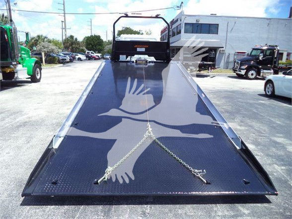 Tow truck flatbed with metal diamond-plate surface and safety chains, faint logo overlay, rear view showing ramp and surrounding lot.