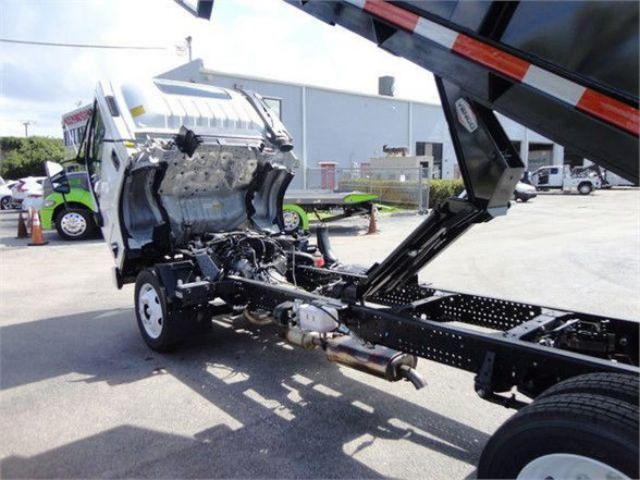 Isuzu NPR HD chassis shown with cab removed, dump truck frame and hydraulic system visible, front wheels, empty engine bay area, partial rear hydraulics.
