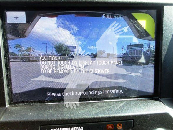 In-vehicle touchscreen display showing reflection of truck exterior with caution text overlaid: 'CAUTION!! DO NOT TOUCH ON DISPLAY TOUCH PANEL DURING INSTALLATION TO BE REMOVED BY THE CUSTOMER' and 'Please check surroundings for safety.'