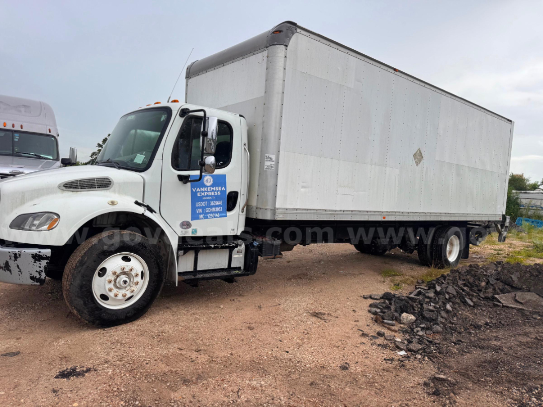 Box truck with white cab and large silver box trailer, blue dealer sticker on door, front wheel visible, on dirt lot.