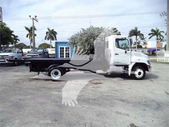 2017 Hino 258LP hooklift truck side view showing cab and flatbed with wheelbase, white cab, black flatbed, dealership lot in background.