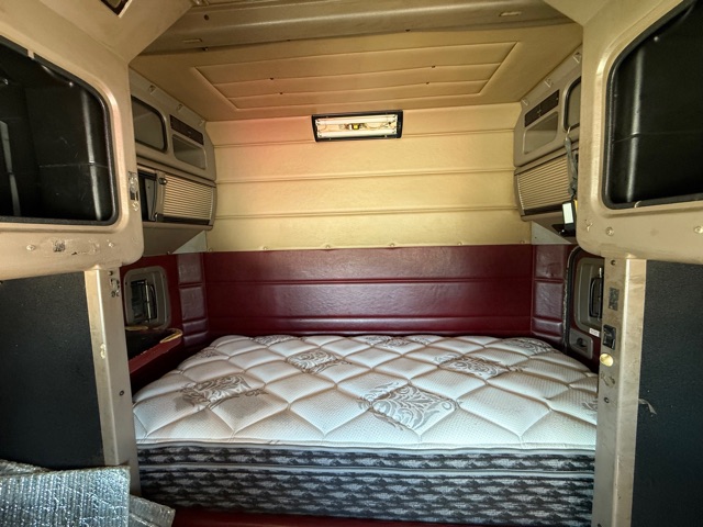 Peterbilt sleeper cab interior with rear-facing bed area, white quilted mattress, cream and tan walls, maroon side panels, and small window.