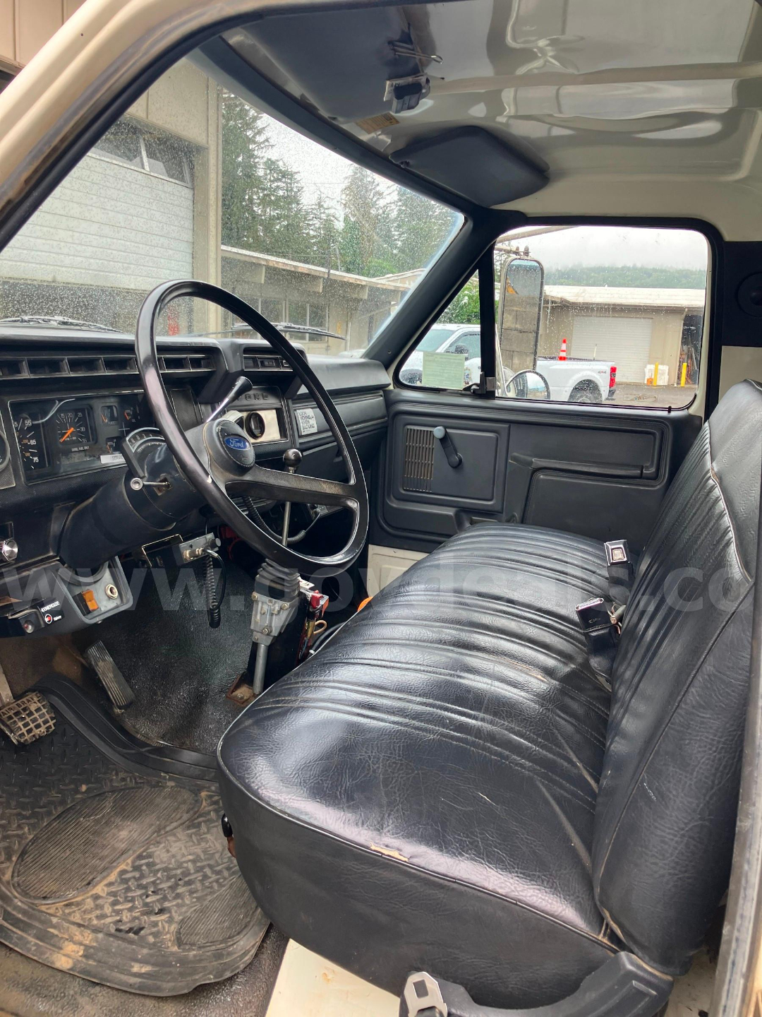 Dashboard and driver’s seat of a 1987 Ford F7000 truck interior showing steering wheel, manual gear lever, vinyl bench seat, and door with pocket. Visible wear on floor mat.