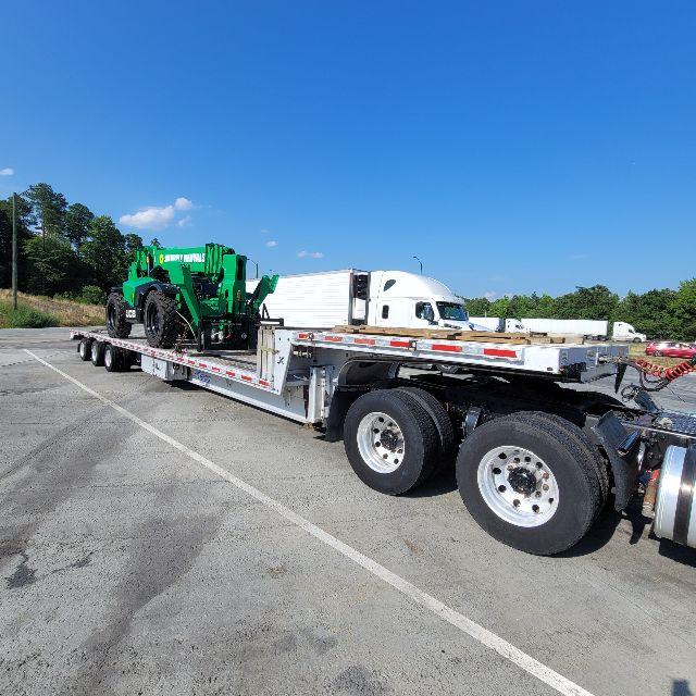 Trailer flatbed showing a green portable machine loaded on a long multi-axle trailer with a white cab in the background.