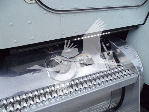 Side view of a 2022 Peterbilt 337 engine bay area showing a polished metal step/rail with perforated pattern and a cylindrical component; part of the truck’s undercarriage.