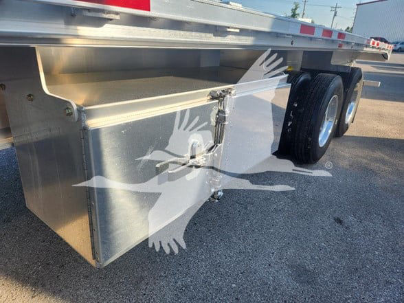 Wabash flatbed trailer showing aluminum toolbox mounted under side with latch, dual rear wheels, and reflective safety tape along the edge.