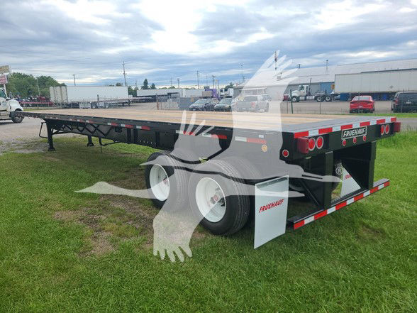 Fruehauf flatbed trailer with dual rear axles, white wheels, and red/white reflective strips. Visible branding 'FRUEHAUF' on the rear bumper and side; on the right rear there are round taillights. The trailer sits on a grassy lot with other