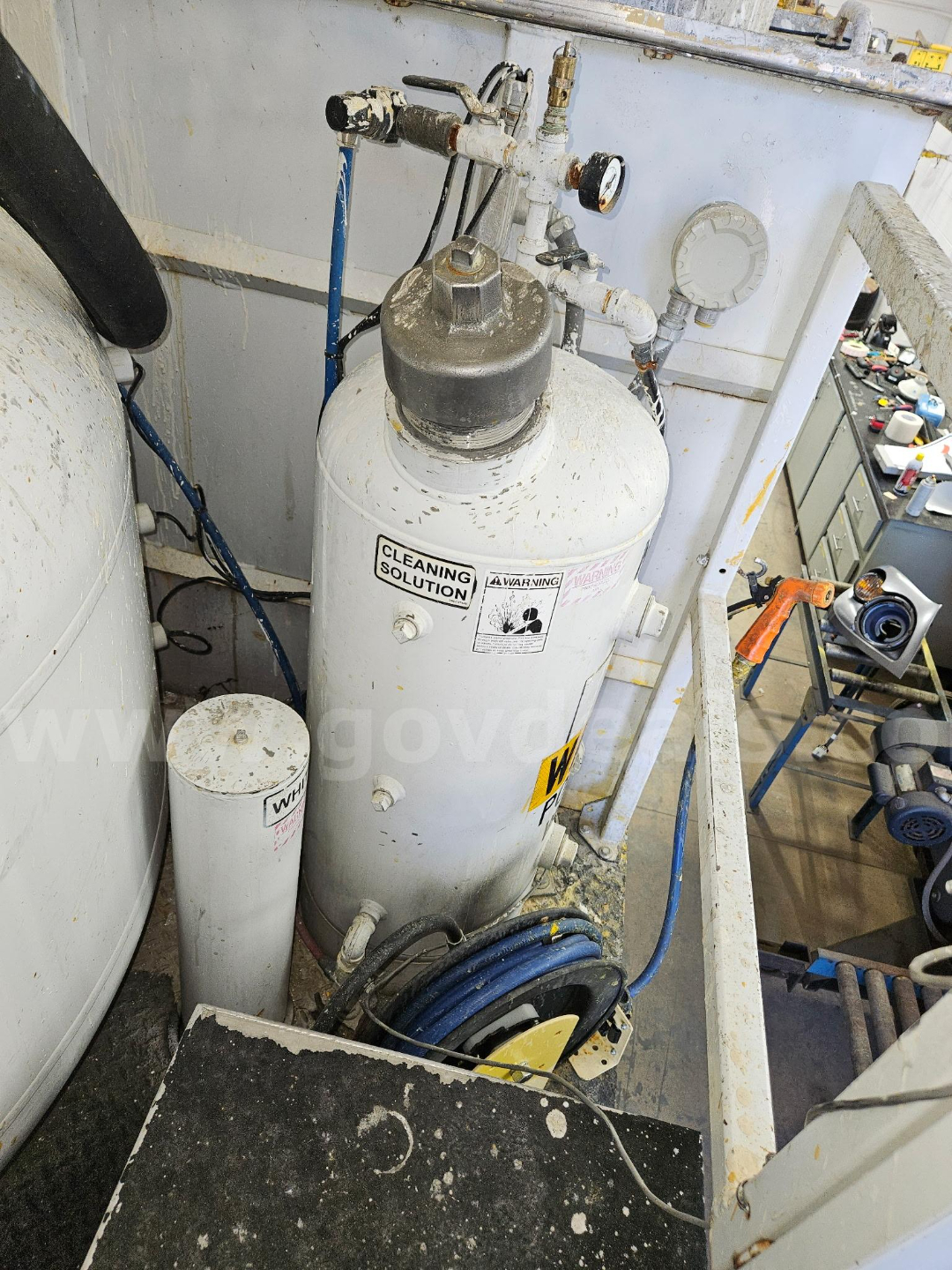 Tank-style air compressor with gauges and piping mounted on a blue frame; label reads CLEANING SOLUTION; gray cylindrical vessels with hoses and controls; various valves and a warning label.