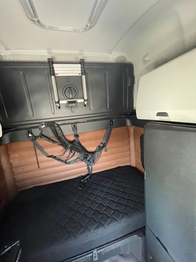 Freightliner Cascadia sleeper cab interior showing bunk area with storage box and netting straps, cabin walls in tan and black, compact cabinet on right side.