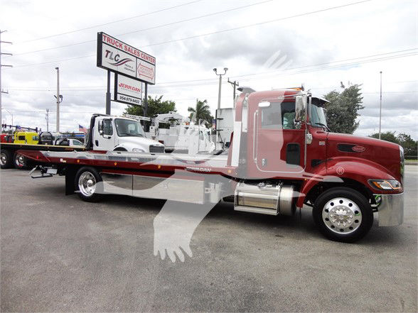 Peterbilt 337 two-axle tow truck flatbed in red with chrome fuel tanks and cab accents; dealership lot in background.