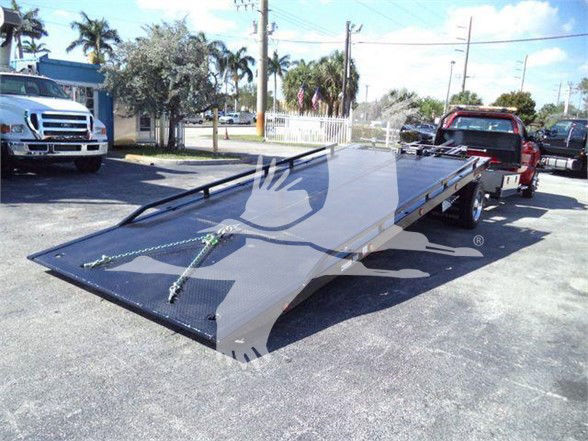 Flatbed tow truck loading ramp with hydraulic mechanism extending from the rear of a Silverado 6500HD chassis, showroom-style lot in background.