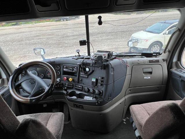 Truck cab interior showing steering wheel, dashboard, and driver area. Various gauges and controls visible on center console; windshield and side mirror view.