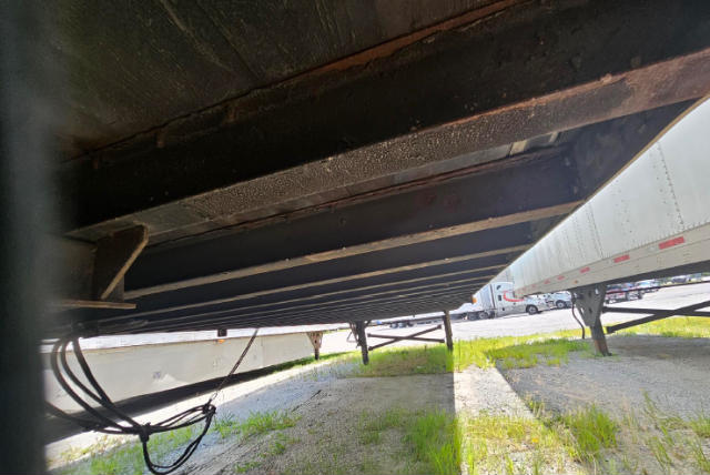 Underneath view of a utility trailer showing frame, support beams, and ground. Visible are hangers, cables, and underside structure.