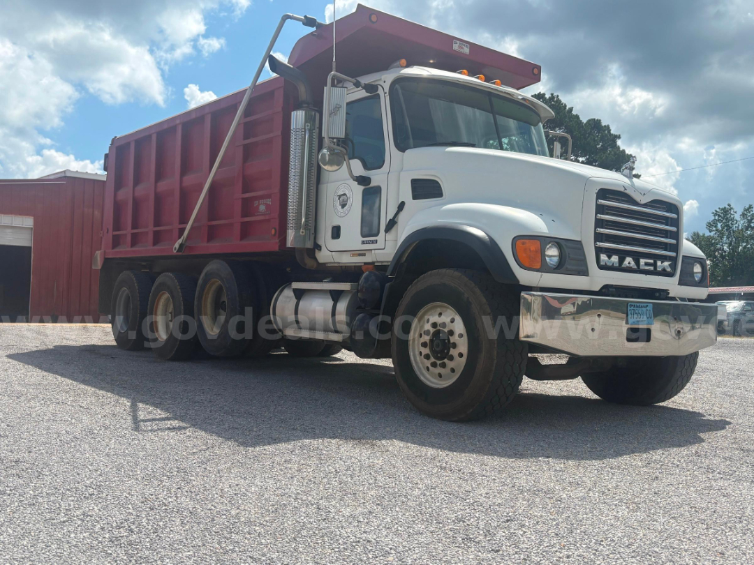 Dump truck with white cab and pink dump body, Mack grille, large tires, side exhaust, parked on gravel.