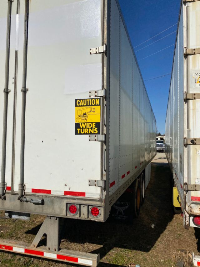 Rear view of a white dry van trailer with 'CAUTION WIDE TURNS' sticker on the door and red reflective strips; tandem wheels visible; blue sky in background.