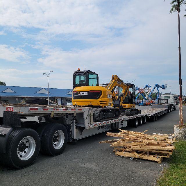 A yellow JCB mini excavator loaded on a long flatbed trailer with stacked lumber in the foreground and a blue sky.