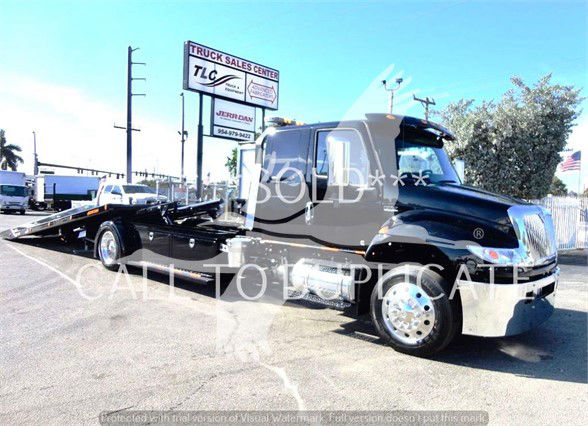 Tow truck with a black cab and a long flatbed in a dealership lot, 'SOLD' watermark across the image, signboards in background.