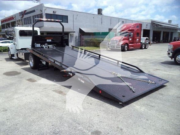 2018 Peterbilt 337 tow truck with a flatbed rollback. The bed is extended, showing latching chains and a diamond plate surface. In the background are service bays and other trucks.