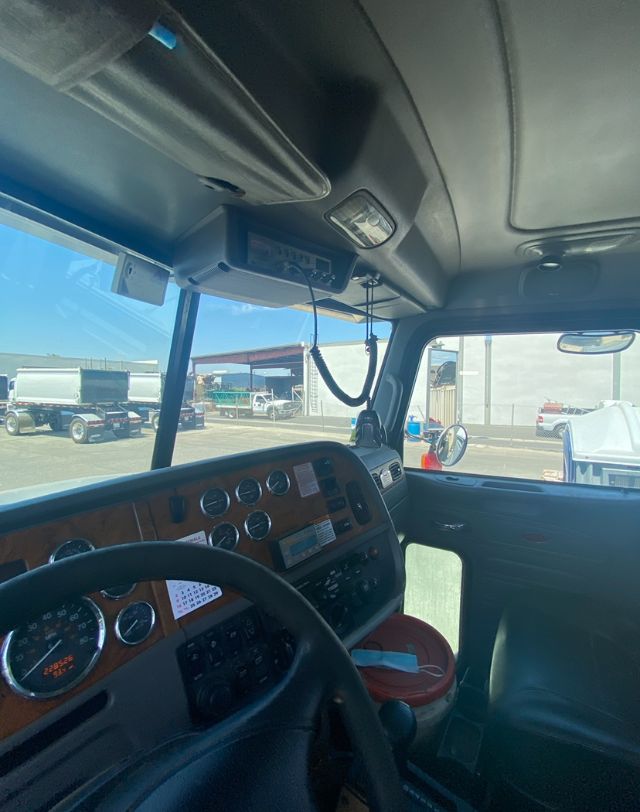 Truck cab interior displaying steering wheel, dashboard with gauges, overhead console, and microphone for the CB radio. Windows show lot with other trucks outside.
