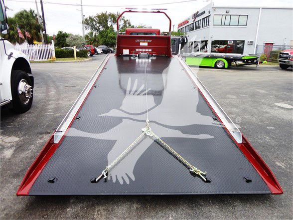 Peterbilt 337 tow truck bed with a dark, textured ramp and two safety chains forming an X, rear view showing hydraulic deck, red cab in background.