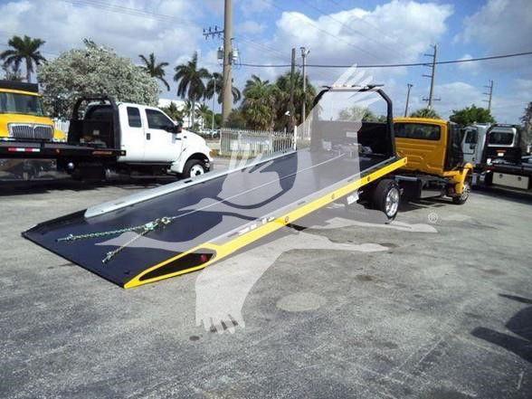 Tow truck with flatbed in an outdoor lot showing a black and yellow ramp and branding on the truck; other trucks and palm trees in background.