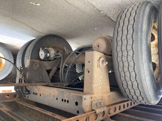 Rear undercarriage of a trailer showing dual axles, tires, leaf spring suspension, and various hoses. Visible components include an axle beam, brake drum, and wiring harness.