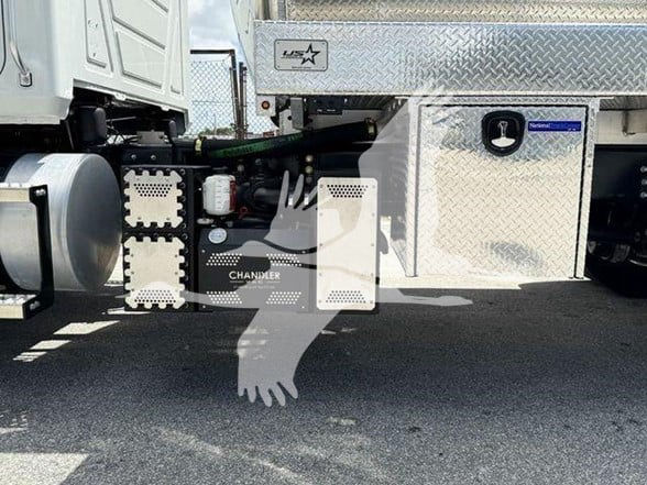 Truck undercarriage showing fuel/air tanks, chassis components, and storage boxes. Visible text includes 'CHAMLER' on a panel and a circular access cap. Diamond plate toolbox and side stickers present.