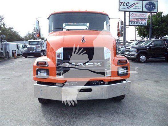 Front view of a bright orange 2024 Mack MD6 tow truck. The grille area shows a large chrome bumper and misty graphic watermark across the center. Headlights at each side, large windshield, side mirrors.