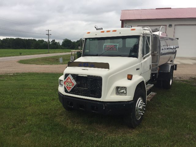 Front view of a white Freightliner FL-70 fueling the truck with a large fuel tank. Hazard placard 1203 on the front bumper. cab features and side steps visible.