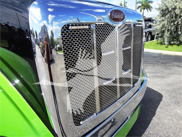 Peterbilt 337 front grille and chrome bumper from a close-up view with green cab and blue sky reflection.