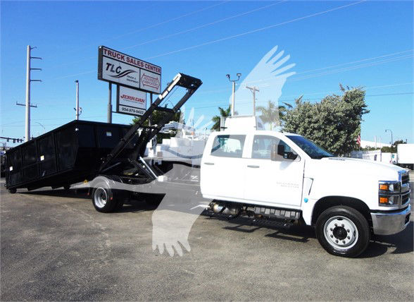 Chevrolet Silverado 5500HD truck with a dump bed in a yard. Visible text on signs includes 'TRUCK SALES CENTER' and 'TLC' on a sign in the background. The truck has a white cab and black dump bed with hydraulic arm.