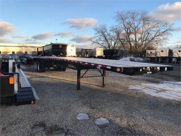 Flatbed trailer (Fontaine Velocity) showing long flat deck with orange reflective tape edge, landing gear, and multiple trailers in background.