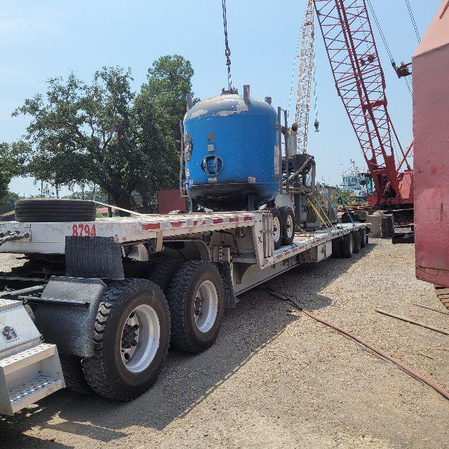 Trailer bed with a blue cylindrical tank on a low-boy trailer, crane in background, gravel surface, trees in distance.