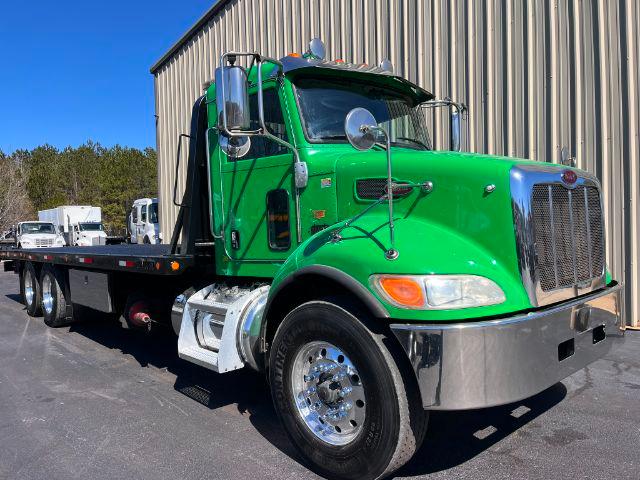 Green Peterbilt 348 rollback truck with long flatbed, chrome grille and dual exhaust mirrors, front wheel and side view; clear weather.