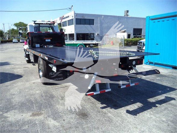 Flatbed tow truck rear with hydraulic bed extended and rear underride guard. Visible light bars on cab, DOT lights, and open machinery area.