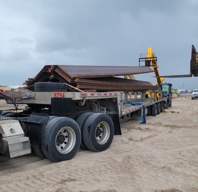 Trailer with stacked steel decking on a flatbed trailer, tires, and a loading crane in a yard.