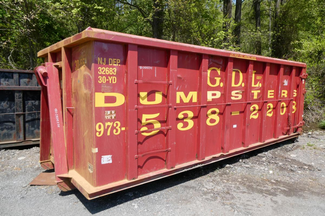 Red garbage or dump dumpster with yellow lettering and numbers, placed outdoors on dirt and gravel, trees in background.