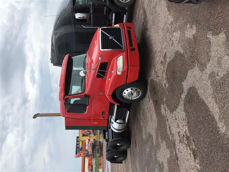 Red Volvo cab-over engine bay and side view showing front grille, chrome exhaust stack, side mirror, and dual rear axles in a truck yard.