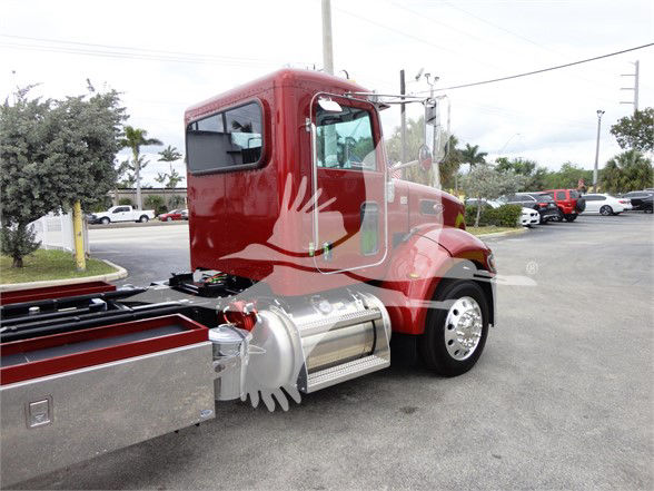 Red 2022 Peterbilt 337 tow truck cab with chrome fuel tanks and side steps, showing rear side and front wheel.
