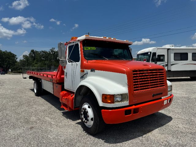 Tow truck with red cab and white fenders, flatbed, International emblem on grille. Windshield has a green and yellow sale sticker. Clear blue sky, other vehicle in background.