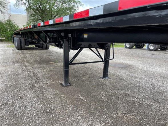 Flatbed trailer with extendable all-steel frame, black undercarriage, supportive legs, and red/white reflective tape along the side. Visible are deck and support legs, with a gravel surface.