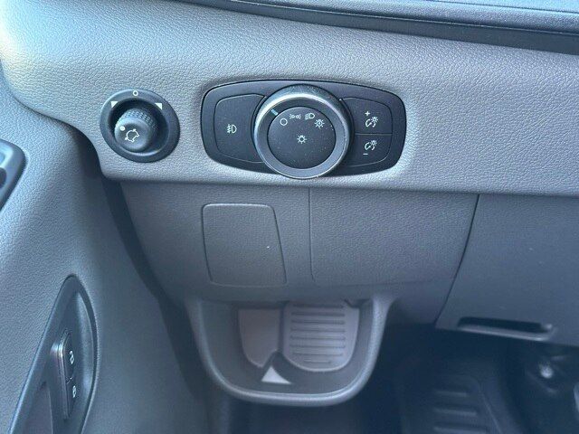 Dashboard left of steering wheel showing headlight control dial with settings and surrounding buttons. Circular dial with symbols for parking lights, headlights, and automatic mode. Left of dial a small switch. A storage pocket below. 