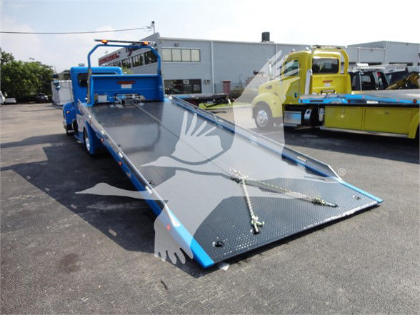 Blue tow truck flatbed loaded with a metallic diamond plate deck and winch lines, parked in lot with other trucks in background.