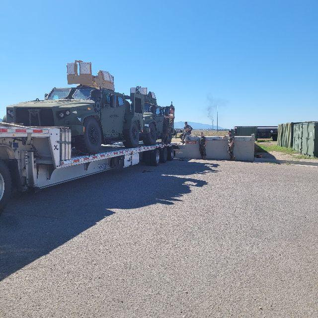 Drop deck trailer carrying military-style armored vehicles loaded on a flatbed trailer with personnel nearby and blue sky.