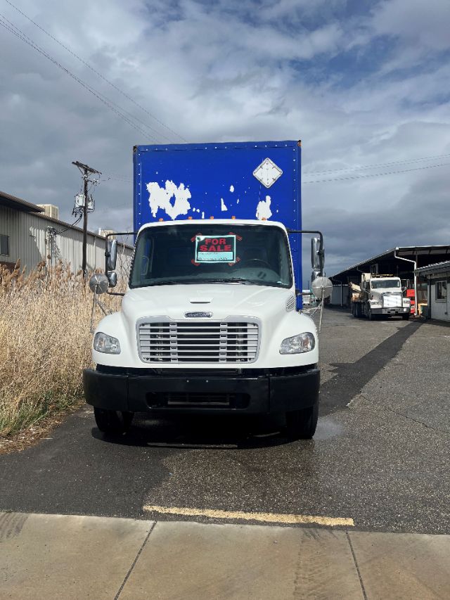 White Freightliner M2 106 box truck front view with a blue box body, 'For Sale' sign on windshield, used vehicle in a lot.