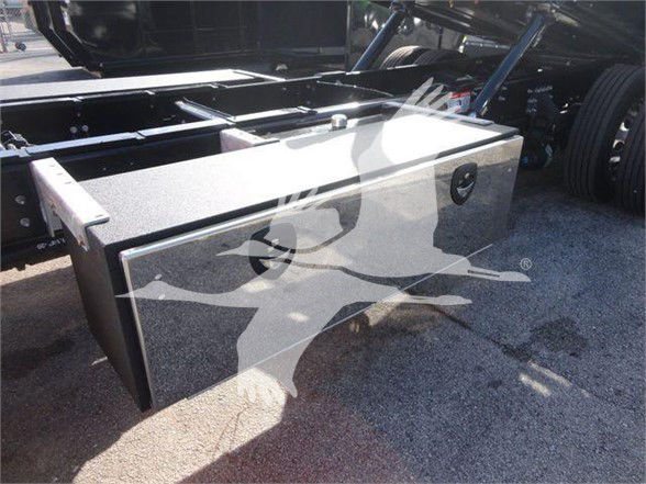 Tow truck chassis showing a large rectangular aluminum toolbox mounted between the cab and front wheel. Visible bolts, textured surface, and two latches.