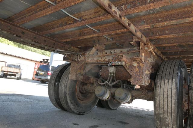 Bottom view of a rusted trailer axle with dual wheels and leaf spring suspension under a flatbed trailer.