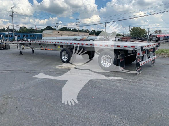 2026 Reitnouer CK-100 flatbed trailer with sliding rear axle, viewed from side rear. Shows long flatbed, dual rear axles, and reflective tape along edges.