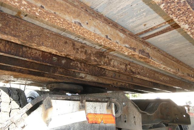 Trailer undercarriage showing rusted crossmembers and frame rails with a small orange reflector and part of the trailer hitch.